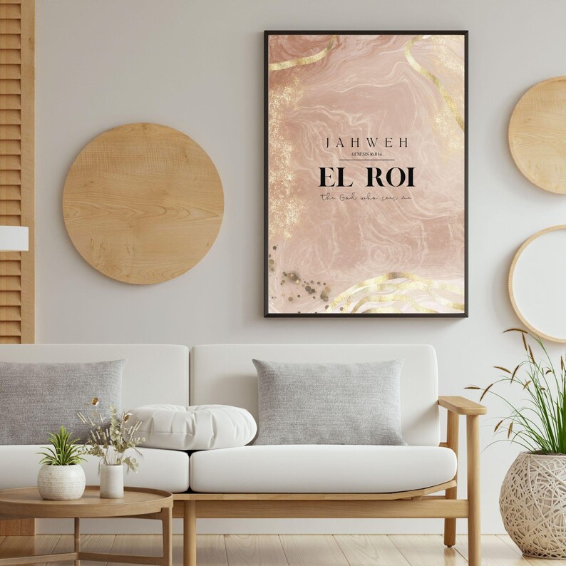 Op de afbeelding: Een ingelijste kunstprint met de tekst "JAHWEH EL ROI the God who sees me" op een roze en gouden abstracte achtergrond. De print hangt boven een witte bank met grijze kussens, aangevuld met houten wanddecoraties en een kleine salontafel.