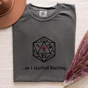 May include: A gray t-shirt featuring a black and red D20 die graphic with the number 1 highlighted in red. Below the graphic, the text reads "...so I started blasting". The shirt is made of a soft material.