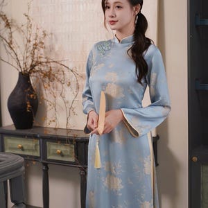 May include: A light blue, long-sleeved dress with a floral pattern. The dress has a mandarin collar and a side slit. A light-colored fan with a yellow tassel is held in the hands. The dress is approximately floor-length.