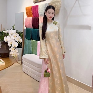 May include: A long-sleeved, floor-length gold tunic with a subtle circular pattern and a green floral brooch at the shoulder, worn over light pink wide-leg trousers. The outfit is paired with a small green drawstring pouch with pearl accents.