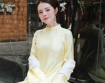 May include: A pale yellow long-sleeved tunic with a high collar and subtle floral embroidery on the shoulder, paired with a white scarf draped over the arms and a white circular handbag adorned with layered circular embellishments.