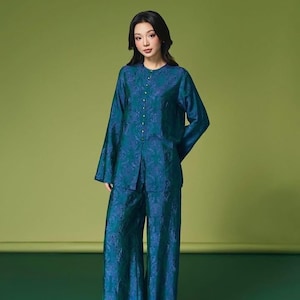 May include: A teal two-piece outfit featuring a long-sleeved top with a button-down front and wide-leg pants. The fabric has a subtle floral pattern. The outfit is worn by a person against a solid green background.