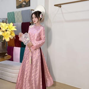 May include: A pink, long-sleeved dress with a floral pattern. The dress has a high neckline and a split skirt, revealing burgundy pants. The person is holding a decorative fan. The background includes colorful fabric samples and a painting.