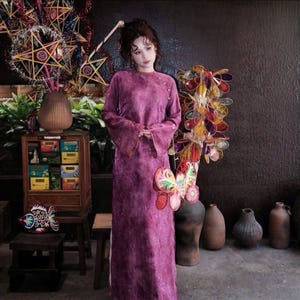 May include: A woman in a long, purple velvet dress stands near a display of colorful lanterns and decorative items. The dress has long sleeves and a high neckline. Various ceramic vases and a wooden cabinet are also visible.