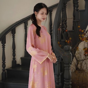 May include: A long, flowing pink dress with gold floral patterns and long sleeves. The dress has a high neckline and a slit on the side. The dress is worn near a dark wooden staircase.