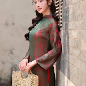 May include: A long-sleeved, green and red patterned dress with a mandarin collar. The dress is paired with a woven straw handbag. The image includes the text "Tạp Hoá USA".