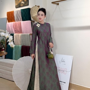 May include: A woman wearing a long, dark green and purple floral patterned dress with a high neckline and long sleeves. The dress has a slit up the side, revealing cream-colored pants. She is holding a small green purse and wearing a flower brooch.