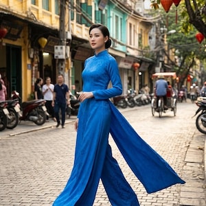 May include: A woman in a vibrant blue traditional Vietnamese dress, the Ao Dai, walks down a cobblestone street. The long-sleeved dress features a mandarin collar and wide, flowing pants. The background shows a bustling street scene with shops and motorbikes.