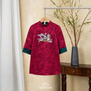 May include: A red dress with a floral pattern and a carousel horse embroidery. The dress has a mandarin collar, three-quarter sleeves with green cuffs, and is hanging on a wooden hanger. The brand name "ahimum" is at the bottom.