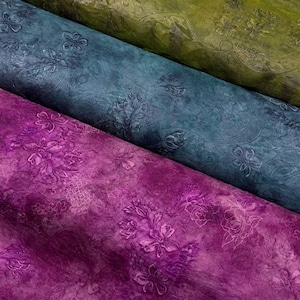 Puede incluir: Primer plano de tres rollos de tela en tonos verdes, turquesa y morado. Cada rollo presenta un estampado floral con detalles en relieve. La tela tiene un aspecto texturizado, con variaciones de color y tono.