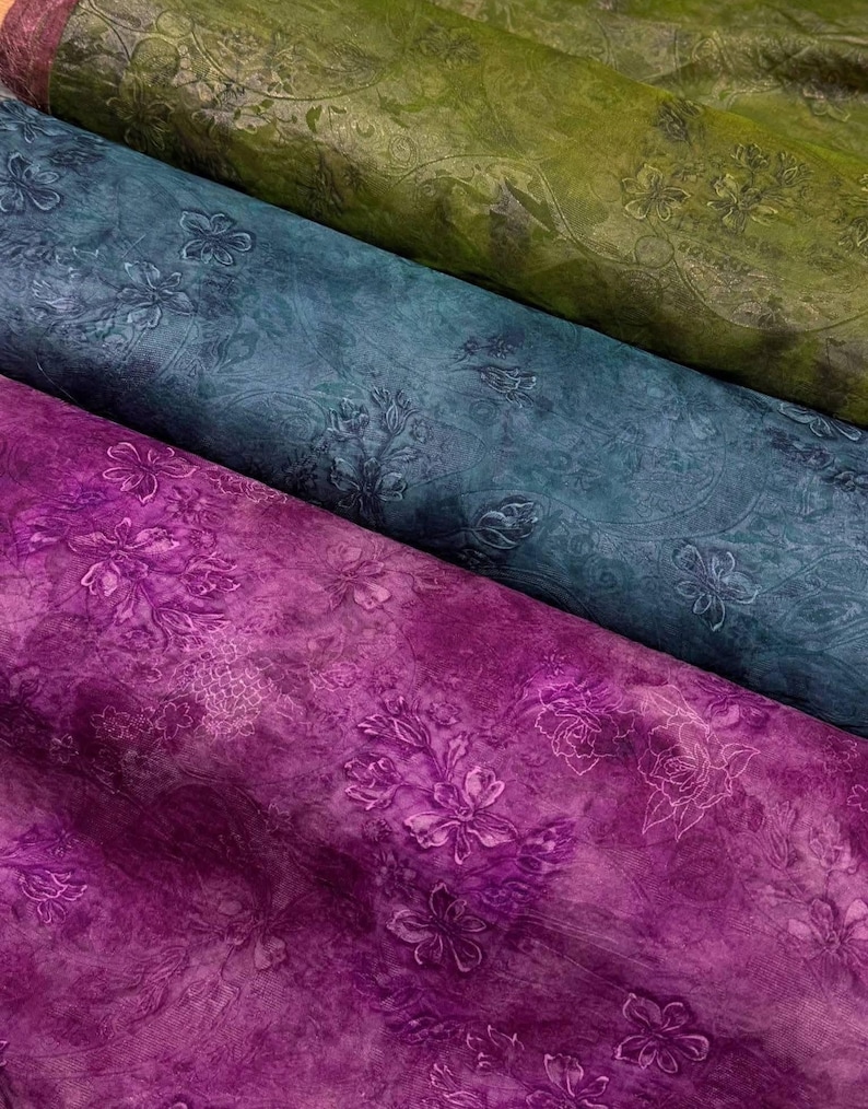 Puede incluir: Primer plano de tres rollos de tela en tonos verdes, turquesa y morado. Cada rollo presenta un estampado floral con detalles en relieve. La tela tiene un aspecto texturizado, con variaciones de color y tono.