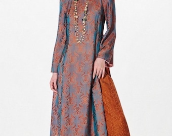 May include: A long, patterned dress in shades of blue, orange, and brown. The dress features a high collar, long sleeves, and a floor-length skirt. A long necklace with multiple strands and beads is worn with the dress. The dress has a side panel in a contrasting orange fabric.