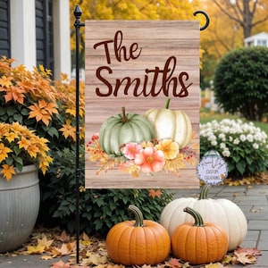 May include: A decorative garden flag with a wooden plank background. The flag features the text "The Smiths" in brown script above a watercolor illustration of pumpkins, flowers, and fall foliage. The flag is displayed on a black metal stand.