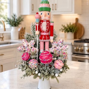 May include: A decorative nutcracker figure in a red and pink uniform stands atop a white base, surrounded by artificial greenery, snowflake-shaped ornaments, and round baubles covered in colorful sprinkles, all arranged in a festive display.