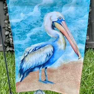 May include: A decorative garden flag featuring a watercolor painting of a pelican standing on a sandy beach. The pelican has blue and white feathers, a long pink beak, and is set against a blue ocean background. The flag has the text "Raini Day Custom Creations".