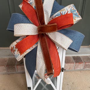 May include: A decorative multi-layered bow featuring a mix of solid navy blue, solid burnt orange, cream with gold polka dots, and cream with a multi-colored leaf pattern ribbon, centered with a vertical strip of brown velvet ribbon.