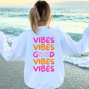Könnte beinhalten: Weißer Pullover mit dem Text "Vibes Vibes Good Vibes Vibes" in rosa und orangefarbener Schrift. Der Pullover wird von einer Person getragen, die an einem Strand steht.