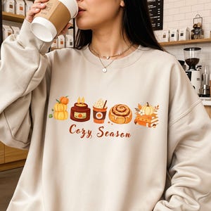 Könnte beinhalten: Ein beigefarbenes Sweatshirt mit Rundhalsausschnitt und einem Grafikdruck von herbstlichen Motiven wie Kürbissen, einer Kerze, einem Kaffeebecher, einer Zimtschnecke und Büchern, darunter der Schriftzug "Cozy Season" in brauner Schreibschrift.