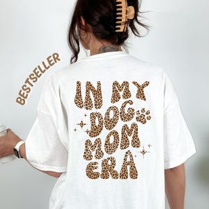 Könnte beinhalten: Weißes T-Shirt mit dem Text "IN MY DOG MOM ERA" in einem Leopardenmuster. Das Wort "BESTSELLER" ist auf der oberen linken Seite des Shirts aufgedruckt. Das Shirt hat einen lässigen Stil.