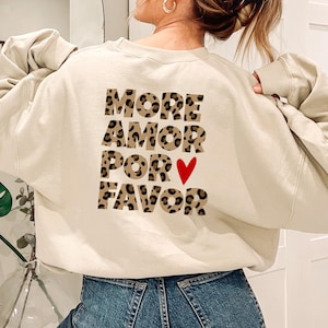 Könnte beinhalten: Cremefarbenes Sweatshirt mit dem Schriftzug "MORE AMOR POR FAVOR" in Leopardenmuster-Buchstaben und einem roten Herz. Das Sweatshirt ist ein lässiger Stil, perfekt für den Alltag.