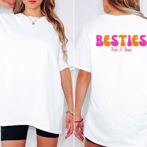 May include: White oversized t-shirt with the word "BESTIES" in pink and orange bubble letters on the back. The names "Lea & Ina" are printed below the word "BESTIES". The shirt is worn with black shorts.