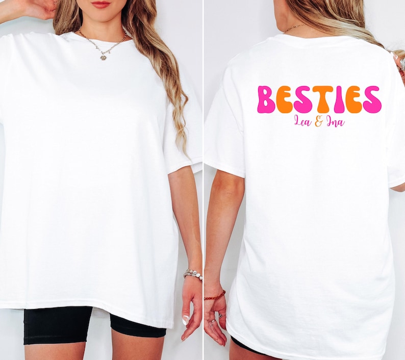 May include: White oversized t-shirt with the word "BESTIES" in pink and orange bubble letters on the back. The names "Lea & Ina" are printed below the word "BESTIES". The shirt is worn with black shorts.