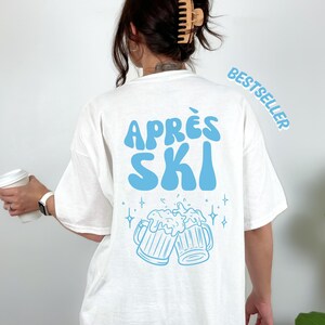 Könnte beinhalten: Ein weißes T-Shirt mit einer hellblauen Grafik auf der Rückseite. Der Text "APRÈS SKI" steht in einer wellenförmigen Schrift über einer Abbildung von zwei anstoßenden Bierkrügen mit Schaum und kleinen Sternen. Das Wort "BESTSELLER" ist zu sehen.