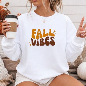 Könnte beinhalten: Ein weißes langärmeliges Sweatshirt mit Rundhalsausschnitt und dem Schriftzug FALL VIBES in einer wellenförmigen Retro-Schriftart. Das Wort FALL ist in Senfgelb und das Wort VIBES in Dunkelbraun gedruckt, akzentuiert durch kleine vierzackige Sterne.