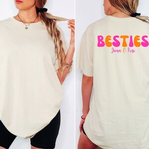 May include: Cream-colored t-shirt with the word "BESTIES" in pink and orange on the back, with the names "Jana & Lisi" below. The shirt is a relaxed fit.