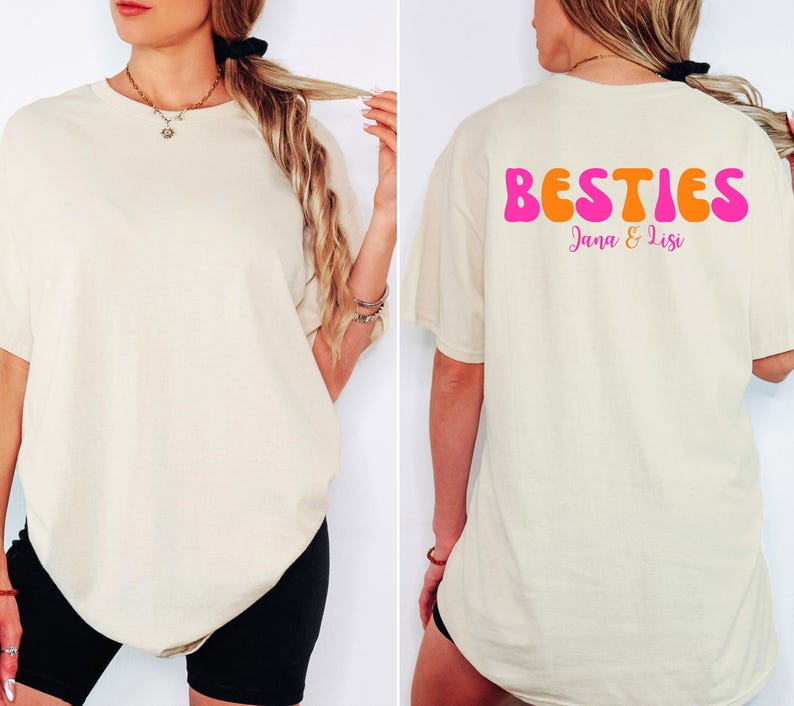 May include: Cream-colored t-shirt with the word "BESTIES" in pink and orange on the back, with the names "Jana & Lisi" below. The shirt is a relaxed fit.
