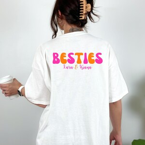 May include: White t-shirt with the word "BESTIES" in pink and orange bubble letters. Below the word "BESTIES" are the names "Lara & Kiana" in a smaller, pink font. The shirt is a casual, oversized style.
