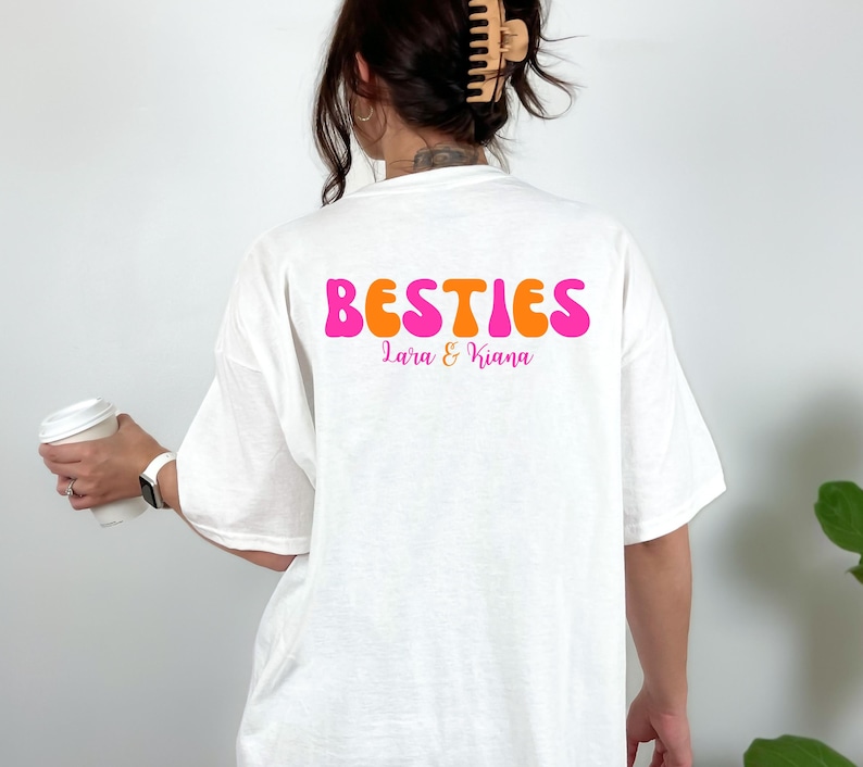 May include: White t-shirt with the word "BESTIES" in pink and orange bubble letters. Below the word "BESTIES" are the names "Lara & Kiana" in a smaller, pink font. The shirt is a casual, oversized style.