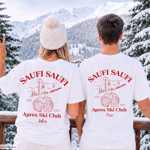 Könnte beinhalten: Zwei weiße T-Shirts mit einem roten Grafikdruck auf der Rückseite. Das Design zeigt eine Berglandschaft mit einem Skilift, zwei anstoßenden Bierkrügen und den Text "SAUFI SAUFI APRES SKI CLUB EST. 2027" mit personalisierten Namen.