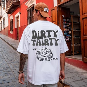 Könnte beinhalten: Ein weißes Kurzarm-T-Shirt mit dem dunkelgrauen Schriftzug "DIRTY THIRTY" auf der Rückseite. Unter dem Text befindet sich eine schwarze Strichzeichnung von zwei anstoßenden Bierkrügen mit Schaum, ergänzt durch "est." und "1996".