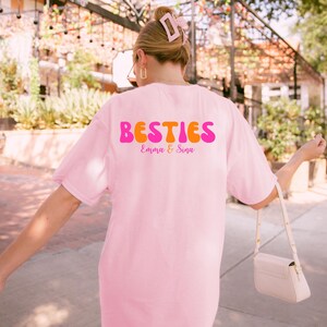 May include: A light pink t-shirt with the word "BESTIES" in a retro font, with the names "Emma & Sina" below. The shirt is a soft pink color.