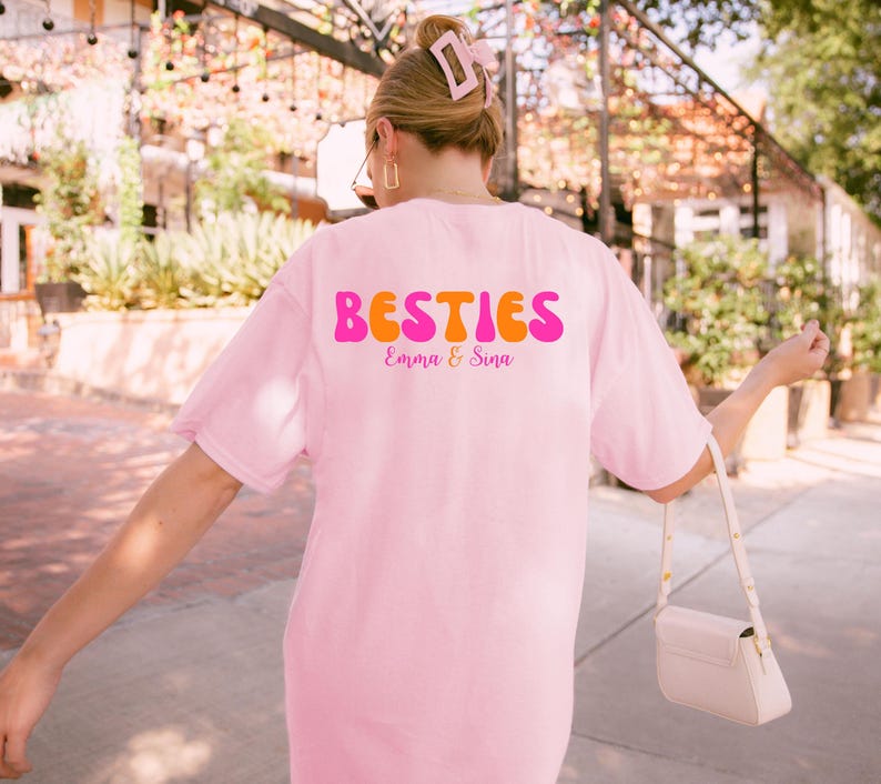 May include: A light pink t-shirt with the word "BESTIES" in a retro font, with the names "Emma & Sina" below. The shirt is a soft pink color.