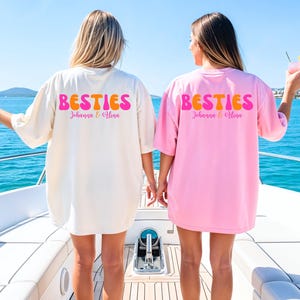 May include: Two oversized t-shirts, one cream and one pink, with the word "BESTIES" in retro orange and pink lettering. The names "Johanna & Alina" are printed below. The shirts are worn on a boat with a blue ocean background.