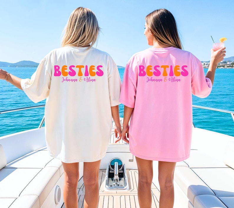 May include: Two oversized t-shirts, one cream and one pink, with the word "BESTIES" in retro orange and pink lettering. The names "Johanna & Alina" are printed below. The shirts are worn on a boat with a blue ocean background.
