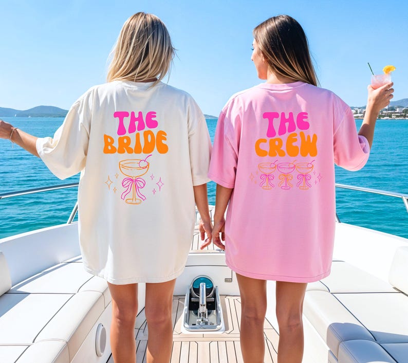 May include: Two oversized t-shirts, one cream and one pink, with retro text and cocktail graphics. The cream shirt says "THE BRIDE" in pink and orange, while the pink shirt says "THE CREW". Both shirts are worn on a boat.