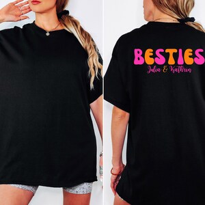 May include: Black t-shirt with the word "BESTIES" in orange and pink bubble letters on the back. Below the word "BESTIES" is the text "Julia & Kathrin" in pink script. The front of the shirt is plain black.