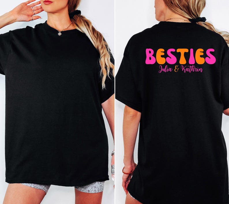 May include: Black t-shirt with the word "BESTIES" in orange and pink bubble letters on the back. Below the word "BESTIES" is the text "Julia & Kathrin" in pink script. The front of the shirt is plain black.