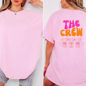 May include: A light pink t-shirt with the text "THE CREW" in pink and orange on the back, along with three cocktail glasses. The front of the shirt is plain pink.