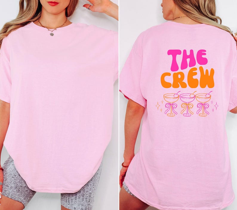May include: A light pink t-shirt with the text "THE CREW" in pink and orange on the back, along with three cocktail glasses. The front of the shirt is plain pink.