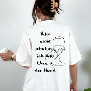 May include: White t-shirt with black text and a wine glass graphic. The text reads "Bitte nicht schubsen ich hab Wein in der Hand". The shirt is a casual style.