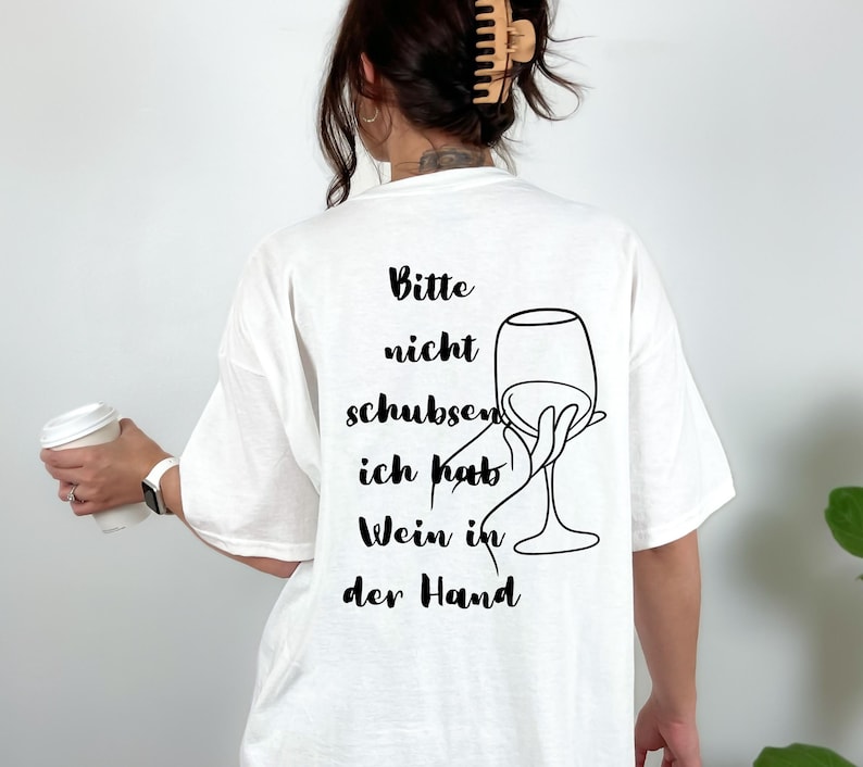 May include: White t-shirt with black text and a wine glass graphic. The text reads "Bitte nicht schubsen ich hab Wein in der Hand". The shirt is a casual style.