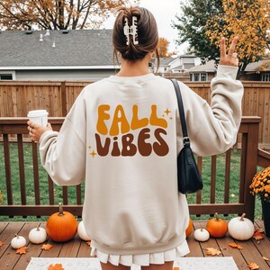 Könnte beinhalten: Ein beiges Langarm-Sweatshirt mit dem Schriftzug FALL VIBES in großen, wellenförmigen orangefarbenen und braunen Buchstaben auf der Rückseite. Das Design enthält kleine Sternakzente. Das Sweatshirt wird mit einem weißen Faltenrock kombiniert.