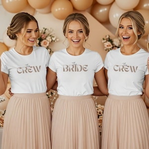 Könnte beinhalten: Drei Frauen tragen weiße T-Shirts mit dem Text "The Crew" und "The Bride" in brauner Schrift. Sie tragen plissierte, hellrosa Röcke und halten Champagnergläser. Ballons sind im Hintergrund.