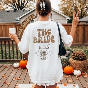 Peut inclure: T-shirt blanc avec l'inscription "THE BRIDE" dans un motif léopard. En dessous, une illustration de verre à cocktail avec une cerise. Le t-shirt fait partie de la collection JGA SHIRTS.
