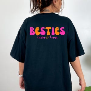 May include: Black t-shirt with the word "BESTIES" in orange and pink, with the names "Laura & Leana" below. The shirt has short sleeves and a relaxed fit.