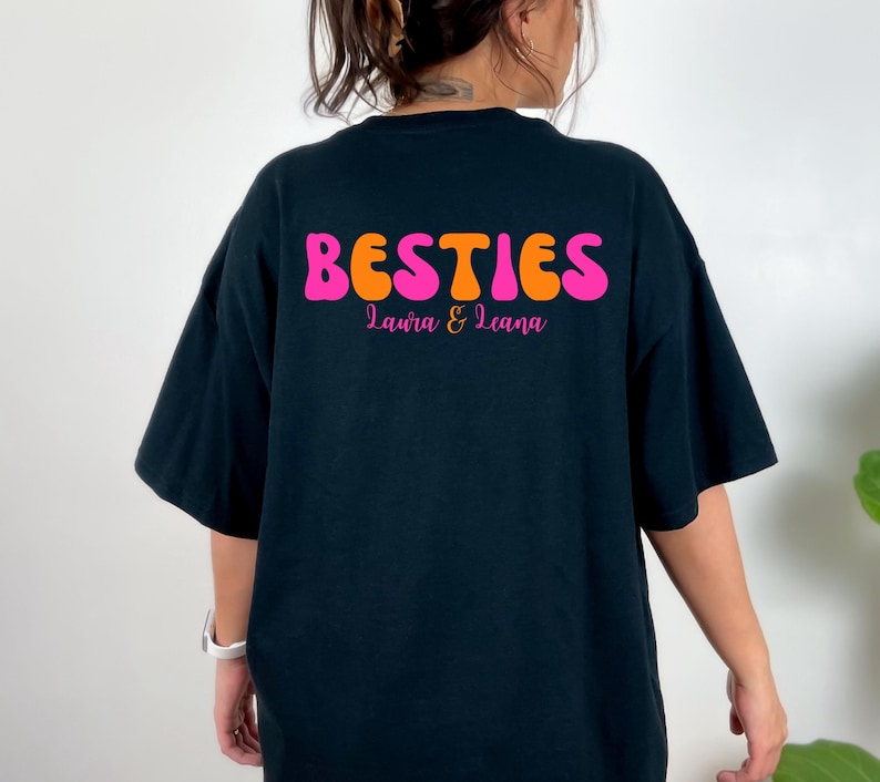 May include: Black t-shirt with the word "BESTIES" in orange and pink, with the names "Laura & Leana" below. The shirt has short sleeves and a relaxed fit.